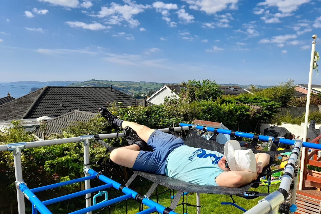 erson relaxing on a cargo net in a garden with a scenic view of the Devon coastline and green rolling hills in the background. They are wearing a SeaNic t-shirt featuring a nature-inspired design, along with a wide-brimmed white sun hat, sunglasses, and outdoor leisurewear. The bright, sunny day highlights the coastal landscape and relaxed lifestyle, embodying SeaNic’s connection to nature, the outdoors, and sustainable clothing